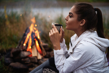 Side view of caucasian female tourist drinking hot tea while sitting near bonfire. Pretty woman...