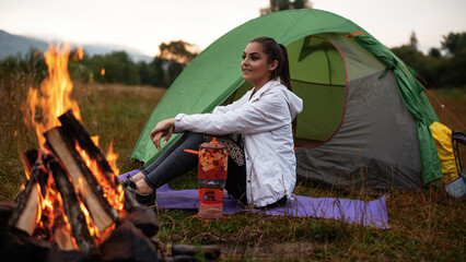 Beautiful female traveler smiling while looking at the nature and resting on grass near bonfire....