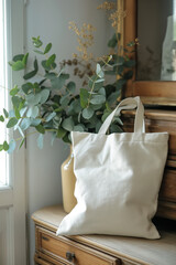 A natural cotton tote bag in an empty white natural cotton tote bag standing on a dresser with a minimalist eucalyptus decoration next to it