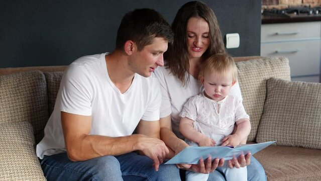 Dad reads book to toddler, child listens while sitting in mom arms, looks at pictures while sitting on couch. Caring parents spend time with adorable baby, learning ,communicating.Happy family concept