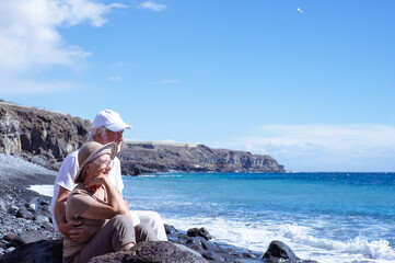 Carefree senior couple in vacation sitting on the seashore looking waves splashing on the beach...