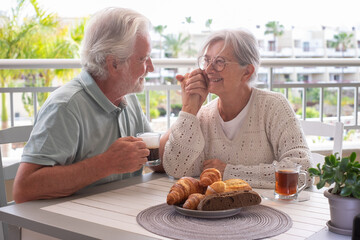 Smiling bonding senior retired couple looking into each other's eyes enjoy breakfast together sitting outdoors on home terrace. Serene retirement lifestyle concept - obrazy, fototapety, plakaty
