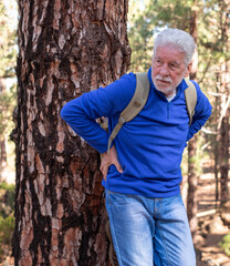 Senior white-haired man hiking in the mountains leans against a tree trunk to catch his breath, touching his back. The elderly pensioner is not feeling very well