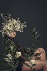 A sensual woman with floral headdress poses amidst lush leaves and blooms in an artistic botanical portrait.