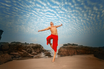 A shirtless man in red pants jumps joyfully against a dramatic sky, with rocky cliffs and a sandy...