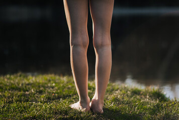 A barefoot girl, a teenage child stands, walks in the summer on a lawn with green grass near the sea, a river in the summer at sunset. Close-up photo of legs in nature.