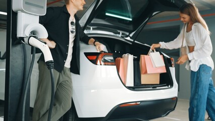 Young couple with shopping bag travel by EV electric car to shopping center parking lot charging in...