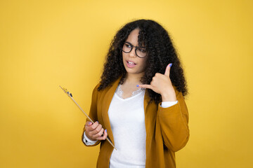 African american business woman with paperwork in hands over yellow background confused doing phone gesture with hand and fingers like talking on the telephone