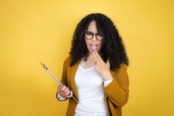 African american business woman with paperwork in hands over yellow background disgusted with her hand inside her mouth