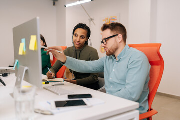 Fototapeta premium Skilled male leader in eyeglasses helping new African female worker with computer corporate software application in office. Motivated young mixed race employees developing project growth strategy