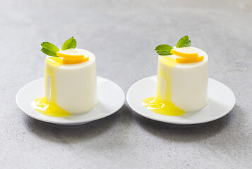 Two desserts. Lemon cream pudding, Panna Cotta cylindrical shape, with lemon sauce, on a plate. Light grey background. Close-up