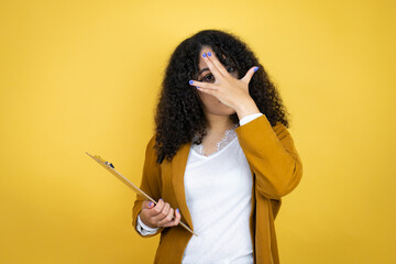 African american business woman with paperwork in hands over yellow background peeking in shock covering face and eyes with hand, looking through fingers with embarrassed expression