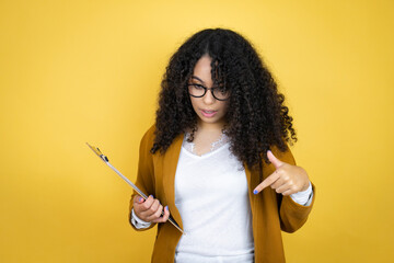 African american business woman with paperwork in hands over yellow background surprised, looking down and pointing down with fingers and raised arms