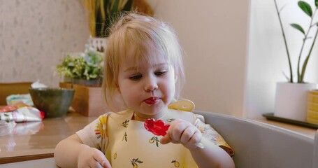 A little girl child toddler eats a lollipop sitting in the kitchen with a bib. 4k video.