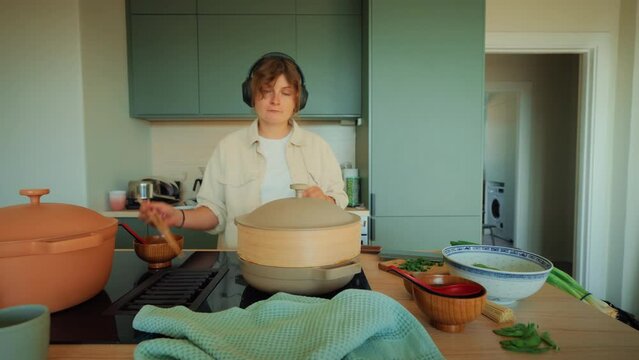 Wide angle shot of young woman cook and try new recipes at home, dance listen to music and taste steamed Asian vegetable and bun dish. Authentic home life of millennials 
