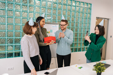 Cheerful attractive African-American woman opening present given by multiethnic colleagues in office to applause of coworkers. Happy team of young business people congratulate woman colleague.