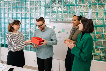Wide shot business team of office employees in festive caps in modern office giving gift to handsome male coworker on birthday. Cheerful man surprised to open box with gift to applause of coworkers. - Powered by Adobe