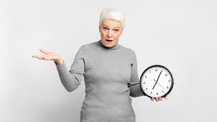 Senior lady surprised holding a clock on grey background