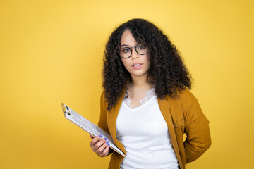 African american business woman with paperwork in hands over yellow background afraid and shocked...