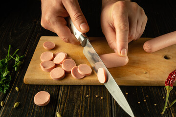 The cook cuts vienna sausage on a wooden cutting board. Preparing delicious sandwiches for...