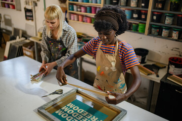 Female interracial graphic workers preparing silkscreen printing plate - obrazy, fototapety, plakaty