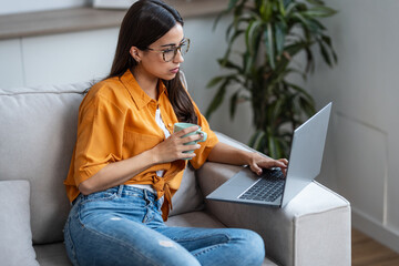 Confident young woman working with her laptop while drinking a cup of coffee sitting on a couch at home