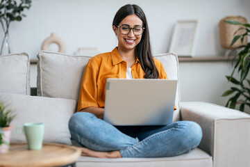 Smiling young woman working with her laptop while sitting on a couch at home