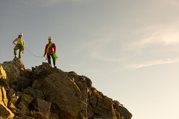 Fototapeta premium Two people are standing on a mountain, one of them wearing a red shirt. The other person is wearing a green harness. The scene is peaceful and serene, with the sun shining brightly in the background