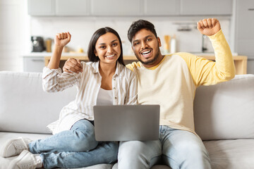 Cheerful Indian couple celebrating at home, using laptop