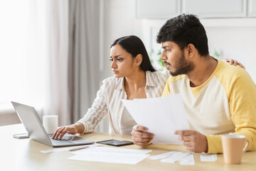 Serious Indian couple discussing finances online, kitchen interior