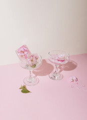 Two glasses with ice cubes and pink flower petals on a pink background. Sunny day concept