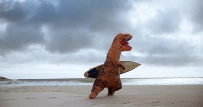 Surfboard, outdoor and dinosaur costume on beach for water sports, competition or training with comedy. Goofy, running and inflatable t rex mascot by ocean or sea for surfing with comic or funny joke