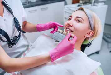 Young female sitting in stomatology clinic chair and smiling showing her teeth. Dentist doctor choosing tooth colors with shade tabs. Healthcare, mouth health and medicare industry concept image