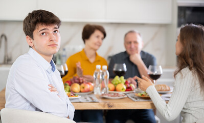 Young guy looking upset and troubled while wife and elderly parents-in-law interacting at table in background during family meal..