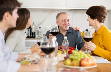 Senior man at kitchen table with wife, grown son and daughter, enjoying conversation at festive table with wine and light snacks
