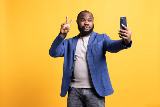 Man using smartphone to take selfies, doing victory hand sign. African american person taking photos using phone selfie camera, showing peace symbol gesturing, studio background