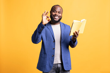 Portrait of smiling man recommending interesting book after being entertained by well written story. Joyous person showing ok hand sign, enjoying literature novel, studio background - obrazy, fototapety, plakaty