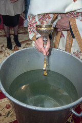 an orthodox priest inserts a cross into the metal baptismal vessel