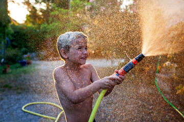 A young boy with joyful laughter sprays water from a garden hose, sparkling droplets flying in the...