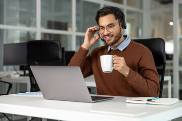 Adult bearded man office employee with headset working on laptop and drinking coffee in office