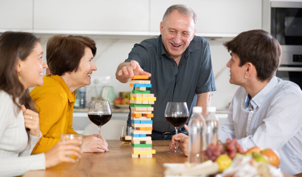 Smiling senior man or father having fun enjoying tower building game with wife and adult children at light comfortable family home