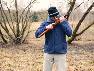 Man with small caliber rifle in nature environment. Outdoor shooting practice to keep reflexes sharp. Male model in blue jacket and dark hat and sunglasses. Dull winter nature background.