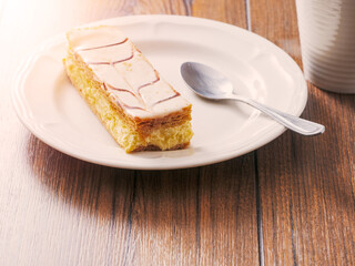 Plate with portion of cram cake on wooden table with spoon and white tea cup. Bakery fine product and dessert. Warm sun flare.