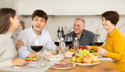 Obraz na płótnie Canvas Smiling young guy engaging in lively conversation with sister and elderly parents at family table with wine and fruits in cozy home kitchen..