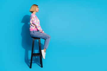 Full length side profile photo of positive girl wear pullover sit on bar chair look at offer empty space isolated on blue color background