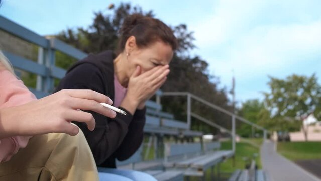 Woman at passive smoking. A view of female feel unwell by smoking girl on the bench outdoor.