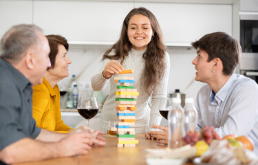 Happy young girl concentrating on removing block from tower while playing jenga, spending free time with boy-friend or husband and parents at light family home - obrazy, fototapety, plakaty