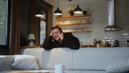  Smiling man in love leaning over the back of the sofa while talking on mobile phone with his...