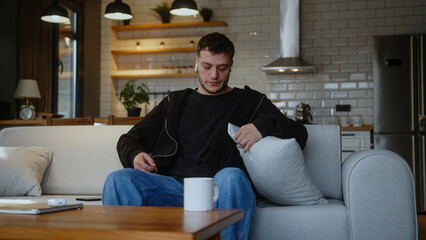 Young man sitting on the sofa and listening to music with wireless in-ear headphones and singing...