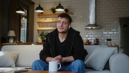 Young adult man sitting on sofa and listening to music with wireless in-ear headphones while drinking coffee or tea from mug. Man having fun, spending time alone at home